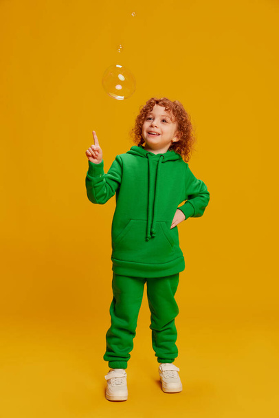 Portrait of cute little girl, child with curly red hair posing, playing with bubbles isolated over yellow background. Concept of childhood, emotions, lifestyle, fashion, happiness. Copy space for ad - Фото, зображення