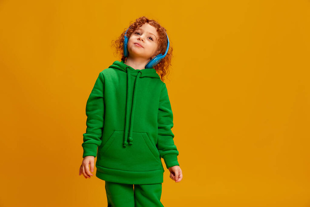 Portrait of cute little girl, child with curly red hair posing in headphones isolated over yellow background. Concept of childhood, emotions, lifestyle, fashion, happiness. Copy space for ad - Photo, image