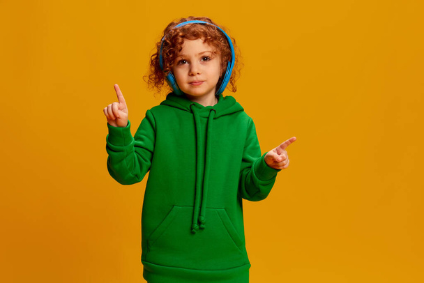 Portrait of cute little girl, child with curly red hair listening to music in headphones isolated on yellow background. Concept of childhood, emotions, lifestyle, fashion, happiness. Copy space for ad - Photo, image