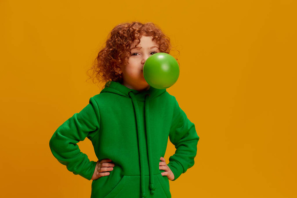 Portrait of cute little girl, child with curly red hair posing with big bubble gum isolated over yellow background. Concept of childhood, emotions, lifestyle, fashion, happiness. Copy space for ad - Фото, изображение