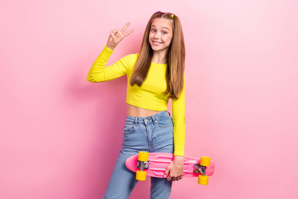 Photo of sweet charming schoolgirl wear yellow crop top riding long board showing v-sign isolated pink color background. - Photo, image