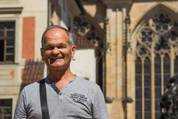 Buon uomo anziano in t-shirt grigia sorridente e guardando la fotocamera sullo sfondo bella architettura. Viaggio turistico anziano nel centro storico di Praga, Repubblica Ceca. Anziani attivi e concetto di viaggio. - Foto, immagini