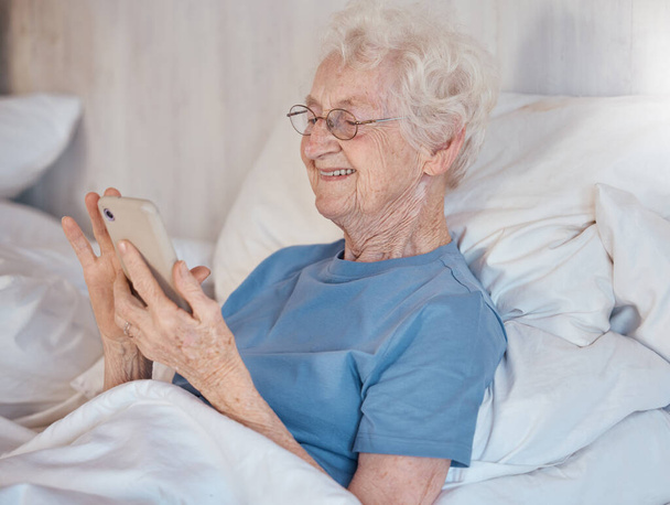 Ηλικιωμένη γυναίκα, σπίτι συνταξιοδότησης και smartphone στο κρεβάτι του γηροκομείου για ιατρική περίθαλψη για να χαλαρώσετε για να υποστηρίξει την υγειονομική περίθαλψη. Πρεσβύτερος με χαρούμενο χαμόγελο, στο κρεβάτι του νοσοκομείου και αποκατάσταση για σωματική υγεία. - Φωτογραφία, εικόνα