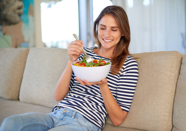 No puedo esperar a morder esta ensalada. una mujer joven comiendo un tazón lleno de ensalada en su sofá en casa - Foto, imagen