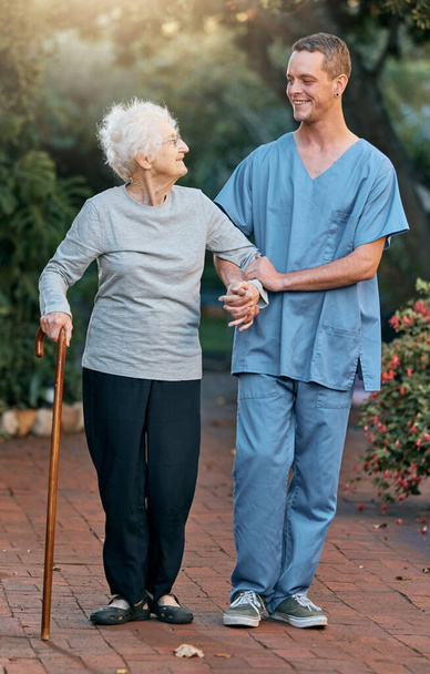 Enfermera, mujer mayor y caminando en el parque para la salud y el bienestar. Fisioterapia, rehabilitación y anciana jubilada con discapacidad y bastón hablando con el médico al aire libre en el jardín - Foto, imagen