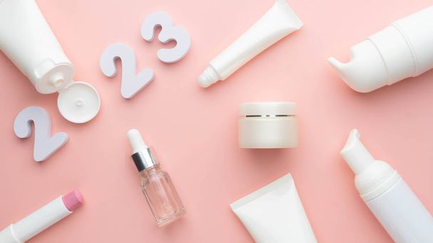 Best skincare products and cosmetic trends of 2023 concept. 2023 white number with lip balm, cream bottle, serum and lotion on pink background. - Foto, imagen