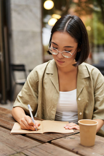 Вертикальный снимок молодой азиатской студентки, девушки в очках, держащей ручку, делающей заметки, пишущей в блокноте и пьющей кофе в кафе. - Фото, изображение