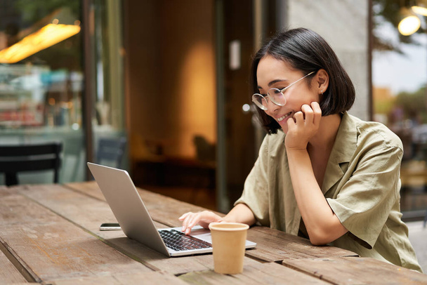 Портрет улыбающейся девушки в очках, сидящей с ноутбуком в открытом кафе, пьющей кофе и работающей удаленно, обучающейся онлайн. - Фото, изображение