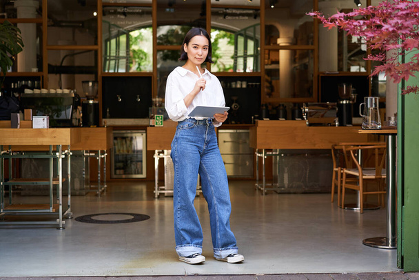 Fiduciosa donna asiatica, cafè manager o dipendente, in piedi vicino all'ingresso con tablet, guardando premuroso. - Foto, immagini