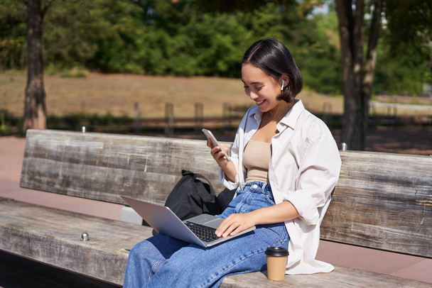 Young asian girl talking on mobile phone, using laptop, working online from park, sitting on bench on sunny day with computer. - Фото, зображення