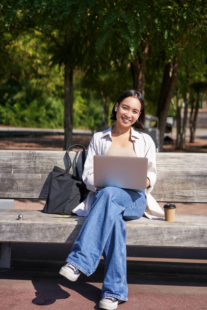 Портрет азиатки, сидящей с ноутбуком на скамейке в парке, слушающей музыку с беспроводными наушниками, делающей домашнее задание, работающей на пульте. - Фото, изображение