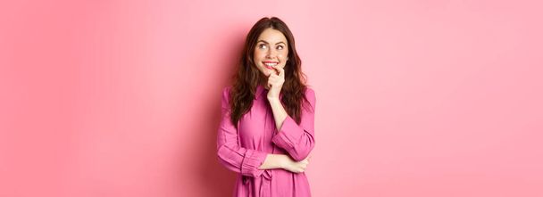 Koketská mladá žena se zamyšleně dívá stranou, usmívá se a kousá do prstu, zatímco přemýšlí, něco si představuje, stojí nad růžovým pozadím. - Fotografie, Obrázek