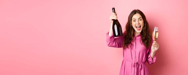 Концепція святкування та свята. Щаслива жінка танцює і розважається, п'є шампанське, тримає пляшку і скло, робить тости, стоїть над рожевим тлом
. - Фото, зображення