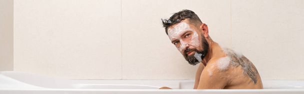 Szakállas férfi maszk az arcon keres Directky a kamera, miközben pihentető fürdőkádban otthon - Fotó, kép