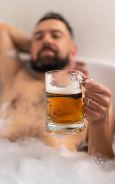 Banyoda bardakta bira tutan adam evde sabunlu, seçici odaklı. - Fotoğraf, Görsel