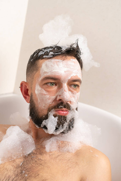 Vicces szakállas férfi fehér maszkkal az arcán és szappan forma a fején otthon fürdik. - Fotó, kép