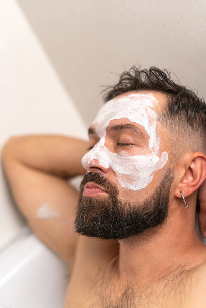 Homme barbu avec masque blanc sur le visage relaxant avec les yeux fermés dans le bain à la maison - Photo, image