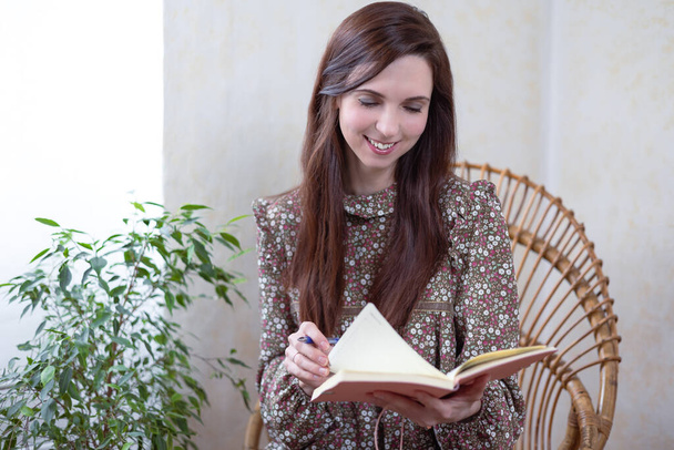 La bella donna sorridente seduta sulla sedia di bambù accanto a un fiore verde e leggendo il suo quaderno. La donna sta lavorando a casa. Lavoro freelance a casa. Una donna che educa a casa. Benessere a casa - Foto, immagini
