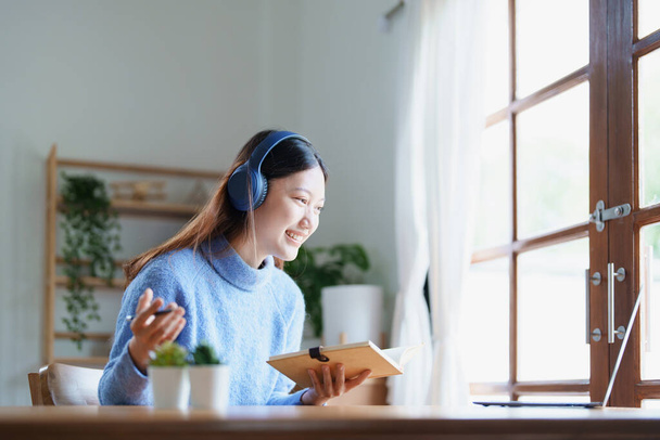 Portrait of a teenage Asian woman using a computer, wearing headphones and using a notebook to study online via video conferencing on a wooden desk at home. - Foto, Imagem