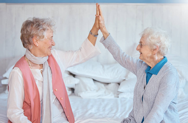 Idős nő, barátok pacsiznak, és mosolyogva ülnek a hálószobában, boldogan vagy együtt az idősek otthonában. Idős nők, legjobb barátok és boldogság támogatással, empátiával és gondoskodással az ágyban a házban. - Fotó, kép