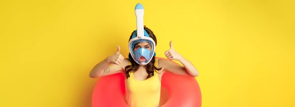 水泳リングで面白い幸せな女性,ダイビングのためのシュノーケリングマスクを身に着けています,親指を表示,良い承認ジェスチャー,黄色の背景に対してポーズ. - 写真・画像