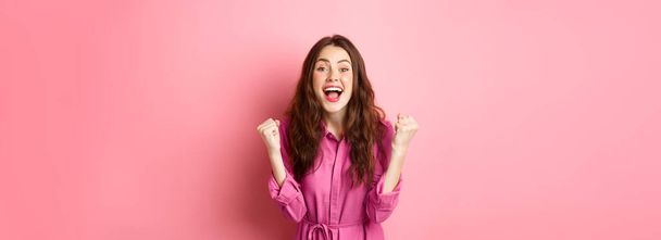 Beautiful smiling woman scream with happy and excited face, saying yes, making fist pump, winning and feeling like champion, triumphing, standing over pink background. - 写真・画像
