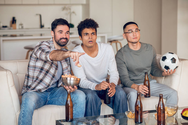 Company of three focused young men sitting on the sofa and watching a football match - Photo, Image