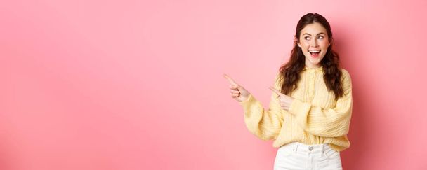Happy good-looking girl demonstrate information, pointing fingers left and staring at promotional text with pleased smile, standing against pink background. - 写真・画像