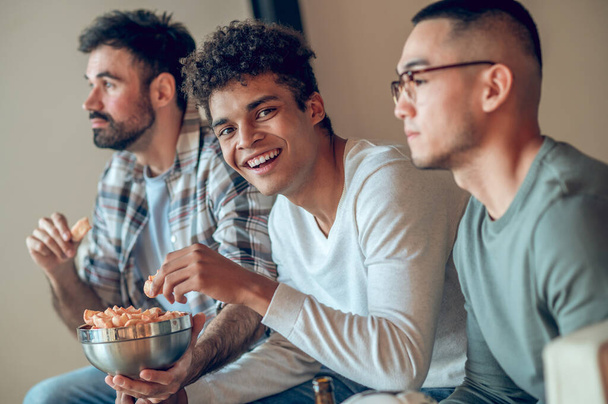 Smiling happy young man eating an appetizer in the company of his calm pensive friends - 写真・画像