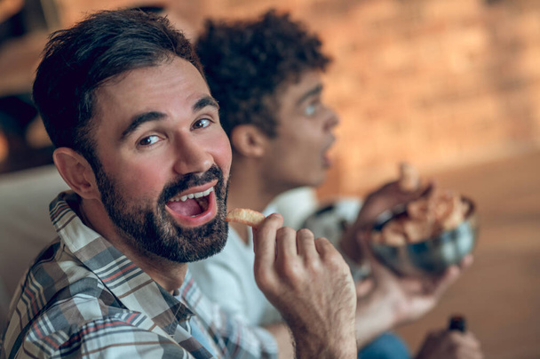 Dark-haired bearded young man eating an appetizer while holding out a bowl of chips to his pal - Foto, afbeelding