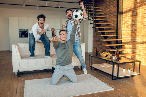 Company of three excited young multiethnic pals screaming and watching a football match on TV - Foto, afbeelding