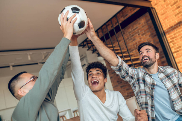 Screaming cheerful man and his two pals lifting together a soccer ball into the air - Foto, immagini