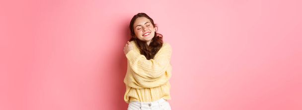 Cute romantic girl falling in love, smiling and hugging herself with dreamy face, standing happy against pink background. - Photo, Image