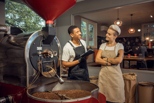 Cheerful young roast master talking to his contented female colleague in a coffee roasting facility - Fotó, kép