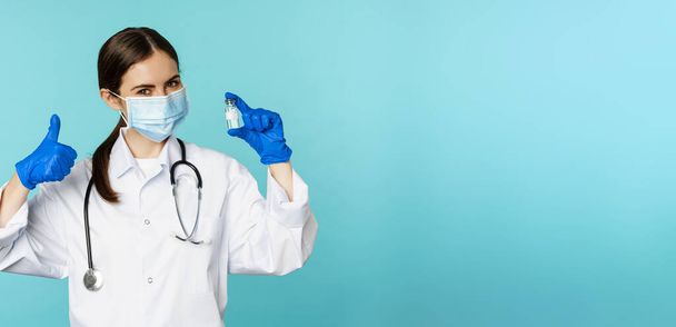 Portrait of smiling doctor, medical personel in face medical mask and rubber gloves, showing thumbs up and coronavirus, omicron vaccine, standing over blue background. - Foto, Bild