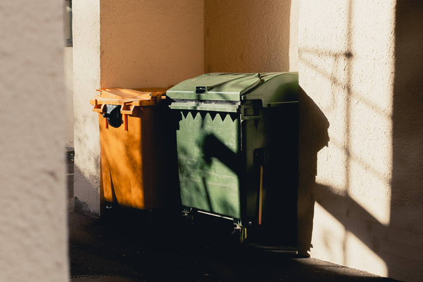 Οι κίτρινοι και πράσινοι κάδοι σκουπιδιών στέκονται στην αψίδα του σπιτιού. Ο ήλιος λάμπει έντονα, σχηματίζοντας παράξενες σκιές.. - Φωτογραφία, εικόνα