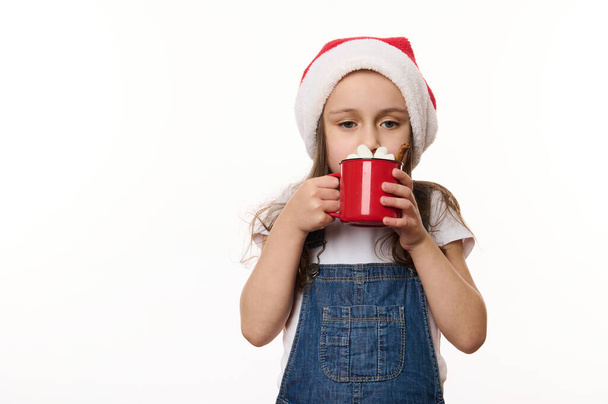 Elszigetelt portré fehér háttér egy aranyos kislány, visel Mikulás kalap, ivás egy finom forró csokoládé mályvacukorral. Varázslatos hangulata a közelgő karácsonyi ünnepek. Ünneplés - Fotó, kép