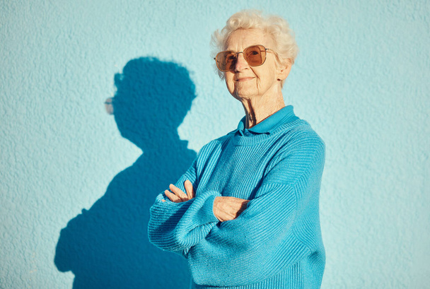 Moda, gafas de sol y retrato de anciana con brazos cruzados sobre fondo de pared para una estética elegante, fresca y única. Retiro, belleza y modelo femenino anciano con gafas de marca de diseño - Foto, imagen