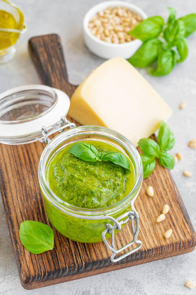 Traditional Italian basil pesto sauce in a glass jar on a board with ingredients for cooking on a gray concrete background. Copy space - Foto, immagini