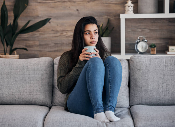 Depression, sad and woman with coffee on a sofa for thinking, lonely and isolated in a living room. Stress, anxiety and girl drinking tea on couch, looking depressed about debt or mental health. - Photo, Image