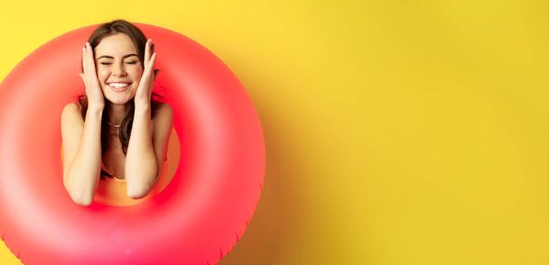 Happy beautiful young woman relaxing on summer vacation, wearing pink swim ring for beach holiday, standing against yellow background. - Foto, imagen