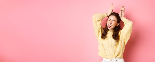 Πορτρέτο του αστείο και χαριτωμένο νεαρή γυναίκα δείχνει γλώσσα, χαμογελώντας χαρούμενος, κάνοντας πασχαλινό λαγουδάκι χειρονομία, στέκεται κατά ροζ φόντο. - Φωτογραφία, εικόνα