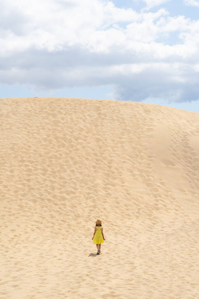 Verticaal landschap met een meisje in een gele jurk in het midden van een berg zand in een woestijn - Foto, afbeelding