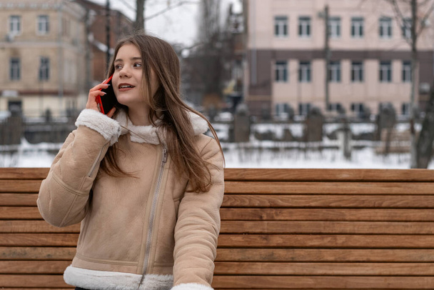 Nuori nainen puhuu puhelimessa ja istuu puistonpenkillä. Tyttö beige lampaan takki lepää ulkona talvella. - Valokuva, kuva
