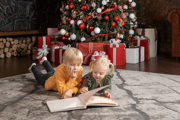 Siblings children on Christmas Eve.Little fair-haired brother and sister read book near the Christmas tree. - Photo, Image