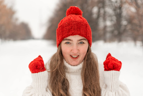 Πορτρέτο του χαρούμενου κοριτσιού σε λευκό μάλλινο πουλόβερ, κόκκινο καπέλο και γάντια σε εξωτερικούς χώρους το χειμώνα. Γυναίκα με ζεστά πλεκτά ρούχα. - Φωτογραφία, εικόνα
