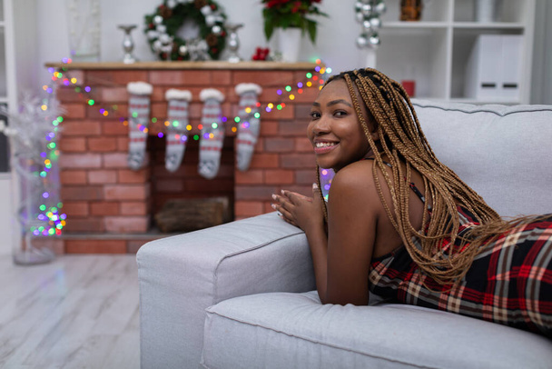 An African woman with braided pigtails lies on a gray sofa. The living room decorated with Christmas decorations. Fireplace with Christmas socks. - Zdjęcie, obraz