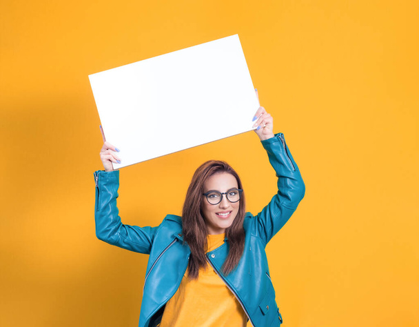 Beautiful women with surprised face holding white speech bubble, wearing blue leather jacket, isolated on yellow background. Female person shows blank empty paper billboard with blank space. Studio shot - Zdjęcie, obraz