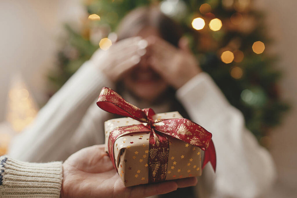Vrolijk kerstfeest en fijne feestdagen! Hand houden van kerst geschenkdoos op de achtergrond van gelukkige vrouw sluiten ogen wachten op verrassing. Cadeaus uitwisselen met kerstboom in sfeervolle ruimte - Foto, afbeelding