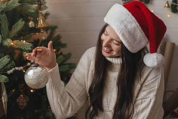 Mutlu Noeller! Rahat süveter ve Noel Baba şapkalı mutlu bir kadın. Atmosferik festival odasında şık takılarla Noel ağacı süslüyor. Kış tatili hazırlığı - Fotoğraf, Görsel